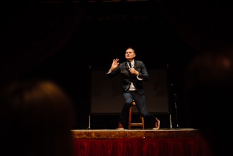 A man is standing on stage holding a microphone, gesturing with one hand while addressing an audience. He is wearing a dark jacket and jeans, and is positioned in front of a large screen. The lighting focuses on him, creating a dramatic atmosphere.