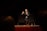 A man is standing on stage holding a microphone, gesturing with one hand while addressing an audience. He is wearing a dark jacket and jeans, and is positioned in front of a large screen. The lighting focuses on him, creating a dramatic atmosphere.