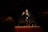 A man is standing on stage holding a microphone, gesturing with one hand while addressing an audience. He is wearing a dark jacket and jeans, and is positioned in front of a large screen. The lighting focuses on him, creating a dramatic atmosphere.