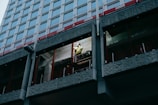 A construction worker wearing a safety helmet and high-visibility vest is standing on scaffolding, working on the exterior of a high-rise building. The building has a modern design with large windows and concrete panels. The worker appears to be focused on a task, possibly involving repairs or installation.