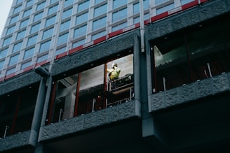 A construction worker wearing a safety helmet and high-visibility vest is standing on scaffolding, working on the exterior of a high-rise building. The building has a modern design with large windows and concrete panels. The worker appears to be focused on a task, possibly involving repairs or installation.