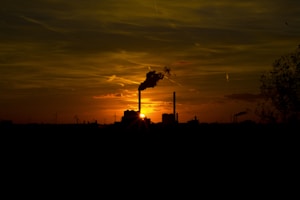 Silhouette of an industrial landscape at sunset with a prominent smokestack emitting smoke. The sky is filled with a warm orange and yellow glow from the setting sun, and wind turbines are visible in the background, providing a contrast between industrial and renewable energy sources. Trees frame the scene on the right.