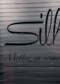 A close-up view of a metallic surface with horizontal ridges. There are large black letters partially visible with more stylized, smaller text beneath that reads 'Mettez en scène'. The colors are subdued with a focus on dark hues against a metallic backdrop.