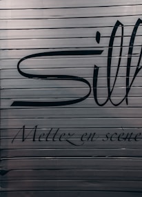 A close-up view of a metallic surface with horizontal ridges. There are large black letters partially visible with more stylized, smaller text beneath that reads 'Mettez en scène'. The colors are subdued with a focus on dark hues against a metallic backdrop.
