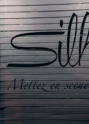 A close-up view of a metallic surface with horizontal ridges. There are large black letters partially visible with more stylized, smaller text beneath that reads 'Mettez en scène'. The colors are subdued with a focus on dark hues against a metallic backdrop.