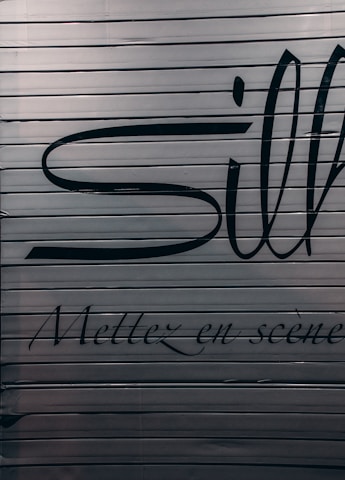 A close-up view of a metallic surface with horizontal ridges. There are large black letters partially visible with more stylized, smaller text beneath that reads 'Mettez en scène'. The colors are subdued with a focus on dark hues against a metallic backdrop.