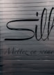 A close-up view of a metallic surface with horizontal ridges. There are large black letters partially visible with more stylized, smaller text beneath that reads 'Mettez en sc&egrave;ne'. The colors are subdued with a focus on dark hues against a metallic backdrop.