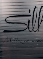A close-up view of a metallic surface with horizontal ridges. There are large black letters partially visible with more stylized, smaller text beneath that reads 'Mettez en sc&egrave;ne'. The colors are subdued with a focus on dark hues against a metallic backdrop.