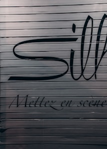 A close-up view of a metallic surface with horizontal ridges. There are large black letters partially visible with more stylized, smaller text beneath that reads 'Mettez en sc&egrave;ne'. The colors are subdued with a focus on dark hues against a metallic backdrop.