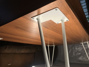A wooden tabletop with a smooth finish is supported by sturdy, white metal legs that are secured with screws. The underside of the table is visible, highlighting the simple and functional design of its construction. The lighting casts soft shadows, enhancing the warm tone of the wood.
