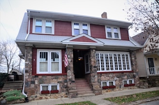 A two-story house with a stone facade and red siding, featuring multiple windows with white frames. An American flag is mounted near the entrance, and there are leafless trees visible around the property.