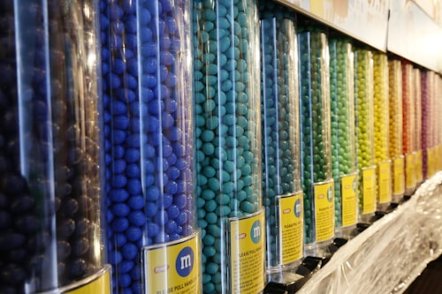 Rows of tall, transparent dispensers are filled with colorful M&M candies, organized by different colors including blue, turquoise, green, yellow, and orange. Each dispenser has a label with an M&M logo and instructions. The arrangement presents a vibrant and orderly display of candies.
