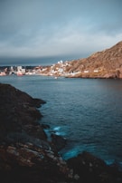 A coastal scene depicting a rocky shoreline on the left leading to a body of water. In the background, a small town with colorful buildings and a few boats in the harbor can be seen on the hillside. The sky is overcast with grey clouds, creating a moody atmosphere.