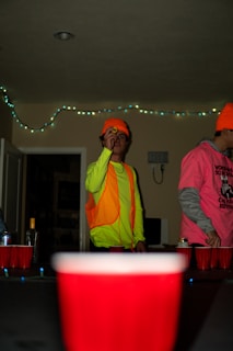 A person wearing a bright neon yellow top and an orange safety vest is holding a ping pong ball, seemingly preparing to throw it. Another individual in a vibrant pink shirt with a graphic design is partially visible beside them. The setting appears to be indoors with dim lighting, and string lights are hanging on the wall in the background. Several red plastic cups are arranged on a table, suggesting a casual party atmosphere.