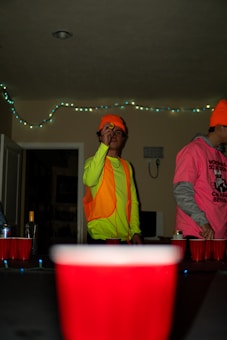 A person wearing a bright neon yellow top and an orange safety vest is holding a ping pong ball, seemingly preparing to throw it. Another individual in a vibrant pink shirt with a graphic design is partially visible beside them. The setting appears to be indoors with dim lighting, and string lights are hanging on the wall in the background. Several red plastic cups are arranged on a table, suggesting a casual party atmosphere.