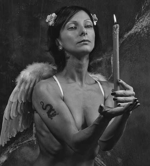 A monochrome photograph depicting a person with short dark hair and flowers as a hair accessory. The individual is wearing angel wings and holds a lit candle upright in both hands. They have a dragon tattoo on their left arm, and their facial expression is serious and contemplative.