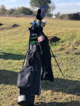 A golf bag stands upright on a grassy field, containing several golf clubs. The golf bag is predominantly black with green accents and features a patch with the number 51. Trees and a partly cloudy sky are visible in the background.