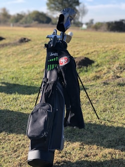 A golf bag stands upright on a grassy field, containing several golf clubs. The golf bag is predominantly black with green accents and features a patch with the number 51. Trees and a partly cloudy sky are visible in the background.