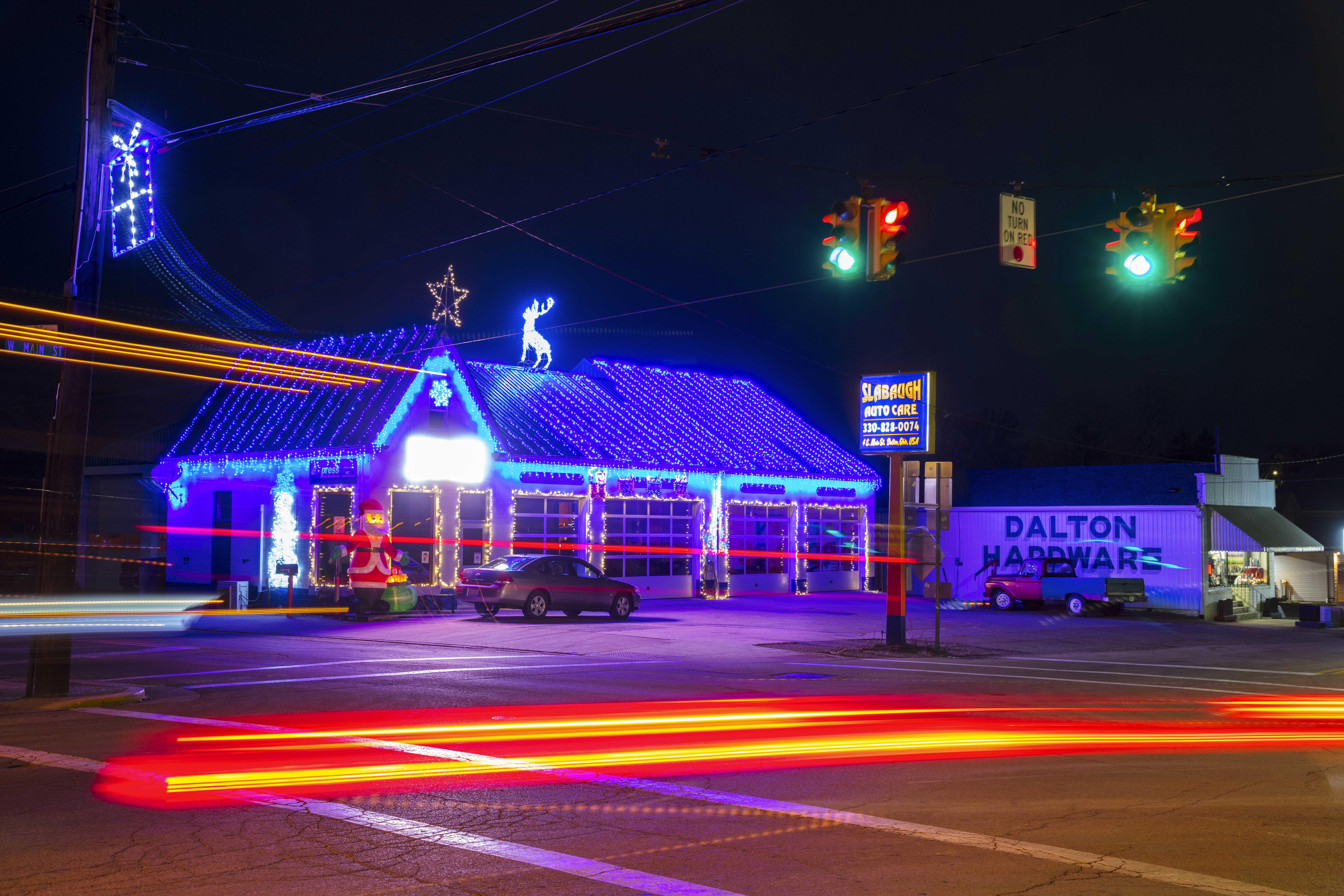 A brightly lit auto care shop adorned with festive blue and white holiday lights, featuring a roof outlined by string lights and adorned with a star and reindeer figures. A Santa Claus inflatable decoration is placed near the entrance. A hardware store with a visible sign stands next to the auto care shop. Traffic lights illuminate the street, and light trails from passing cars create vibrant streaks across the image.