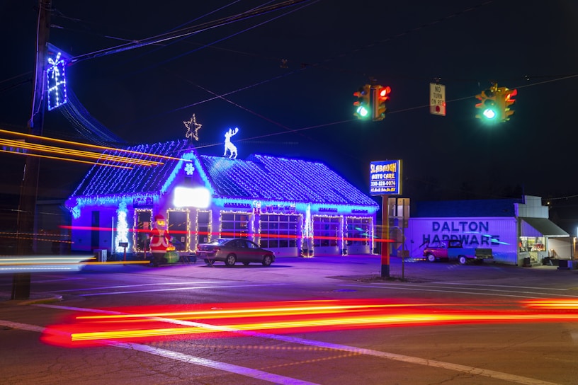 A brightly lit auto care shop adorned with festive blue and white holiday lights, featuring a roof outlined by string lights and adorned with a star and reindeer figures. A Santa Claus inflatable decoration is placed near the entrance. A hardware store with a visible sign stands next to the auto care shop. Traffic lights illuminate the street, and light trails from passing cars create vibrant streaks across the image.