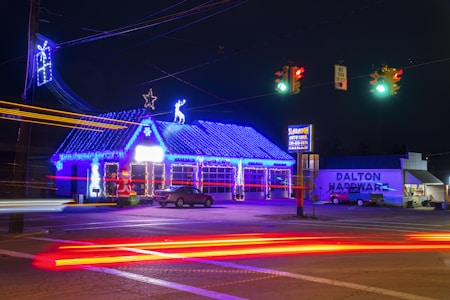 A brightly lit auto care shop adorned with festive blue and white holiday lights, featuring a roof outlined by string lights and adorned with a star and reindeer figures. A Santa Claus inflatable decoration is placed near the entrance. A hardware store with a visible sign stands next to the auto care shop. Traffic lights illuminate the street, and light trails from passing cars create vibrant streaks across the image.
