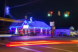 A brightly lit auto care shop adorned with festive blue and white holiday lights, featuring a roof outlined by string lights and adorned with a star and reindeer figures. A Santa Claus inflatable decoration is placed near the entrance. A hardware store with a visible sign stands next to the auto care shop. Traffic lights illuminate the street, and light trails from passing cars create vibrant streaks across the image.