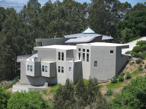 A modern, angular house with a gray facade is nestled on a hillside surrounded by dense green foliage. The structure features multiple levels and geometric designs, including numerous large windows and a small rooftop terrace. The surrounding landscape includes lush trees and greenery, suggesting the house is located in a forested or suburban area.
