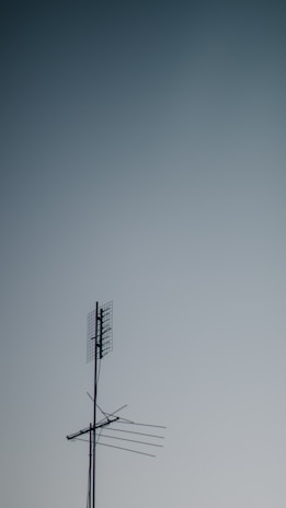 A metallic television antenna with several horizontal rods and a mesh grid atop extends against a gradient sky background that transitions from light to darker blue.