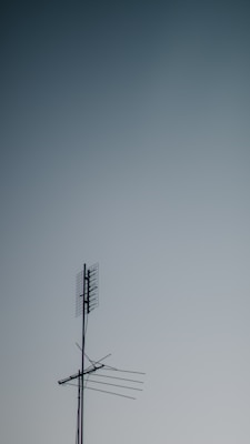 A metallic television antenna with several horizontal rods and a mesh grid atop extends against a gradient sky background that transitions from light to darker blue.