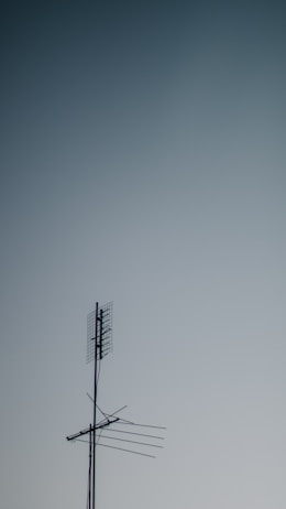 A metallic television antenna with several horizontal rods and a mesh grid atop extends against a gradient sky background that transitions from light to darker blue.
