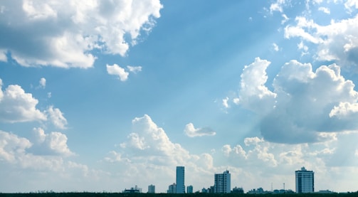 A clear, expansive sky filled with fluffy clouds above a city skyline. Tall buildings rise in the distance, silhouetted against the bright, blue sky. The image conveys a sense of openness and space.