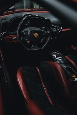The interior of a luxury sports car featuring a sleek steering wheel with a prominent yellow logo, wrapped in black leather. The dashboard is designed with elegant lines and smooth surfaces, incorporating a high-tech appearance. The seating is upholstered in a combination of black and deep red leather, giving the cabin a classic yet modern look.