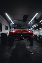 A red sports car is seen elevated on a hydraulic lift inside a dimly lit garage. The lighting creates a dramatic ambiance, highlighting the vehicle's sleek design. Fluorescent lights overhead provide a cool contrast to the dark surroundings. Various automotive equipment and tools can be seen scattered around the workshop.
