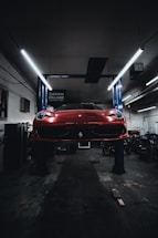 A red sports car is seen elevated on a hydraulic lift inside a dimly lit garage. The lighting creates a dramatic ambiance, highlighting the vehicle's sleek design. Fluorescent lights overhead provide a cool contrast to the dark surroundings. Various automotive equipment and tools can be seen scattered around the workshop.
