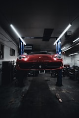 A red sports car is seen elevated on a hydraulic lift inside a dimly lit garage. The lighting creates a dramatic ambiance, highlighting the vehicle's sleek design. Fluorescent lights overhead provide a cool contrast to the dark surroundings. Various automotive equipment and tools can be seen scattered around the workshop.