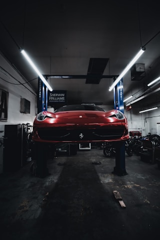 A red sports car is seen elevated on a hydraulic lift inside a dimly lit garage. The lighting creates a dramatic ambiance, highlighting the vehicle's sleek design. Fluorescent lights overhead provide a cool contrast to the dark surroundings. Various automotive equipment and tools can be seen scattered around the workshop.