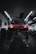 A red sports car is seen elevated on a hydraulic lift inside a dimly lit garage. The lighting creates a dramatic ambiance, highlighting the vehicle's sleek design. Fluorescent lights overhead provide a cool contrast to the dark surroundings. Various automotive equipment and tools can be seen scattered around the workshop.