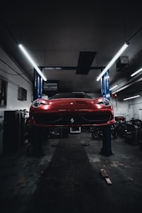 A red sports car is seen elevated on a hydraulic lift inside a dimly lit garage. The lighting creates a dramatic ambiance, highlighting the vehicle's sleek design. Fluorescent lights overhead provide a cool contrast to the dark surroundings. Various automotive equipment and tools can be seen scattered around the workshop.