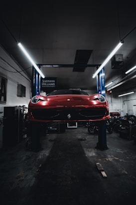 A red sports car is seen elevated on a hydraulic lift inside a dimly lit garage. The lighting creates a dramatic ambiance, highlighting the vehicle's sleek design. Fluorescent lights overhead provide a cool contrast to the dark surroundings. Various automotive equipment and tools can be seen scattered around the workshop.