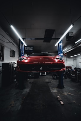 A red sports car is seen elevated on a hydraulic lift inside a dimly lit garage. The lighting creates a dramatic ambiance, highlighting the vehicle's sleek design. Fluorescent lights overhead provide a cool contrast to the dark surroundings. Various automotive equipment and tools can be seen scattered around the workshop.
