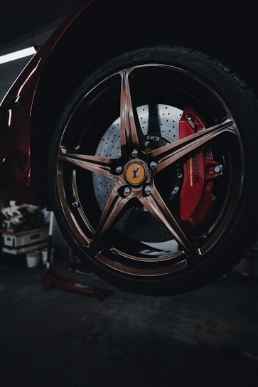 A close-up view of a well-polished car wheel, featuring a bronze-colored alloy rim and a red brake caliper, with a distinctive emblem at the center of the wheel. The background suggests a dimly lit garage or workshop setting.