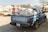 A blue pickup truck with a NISSAN logo is parked outdoors, loaded with large sacks secured in the back. There are buildings and other vehicles in the background, with trees offering partial shade.