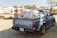 A blue pickup truck with a NISSAN logo is parked outdoors, loaded with large sacks secured in the back. There are buildings and other vehicles in the background, with trees offering partial shade.