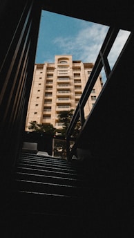 A tall residential building viewed from below through a dark passage or alley. The structure is illuminated by natural sunlight, highlighting the light beige exterior against a partly cloudy blue sky. Shadows create a dramatic contrast between the bright building and the dark foreground.