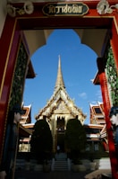 A grand, ornate temple entrance with a detailed archway leads to a glittering, golden temple structure. Intricate architectural details and lush greenery, including two large trees, are visible in the foreground. Vibrant colors and traditional design elements characterize the temple complex.