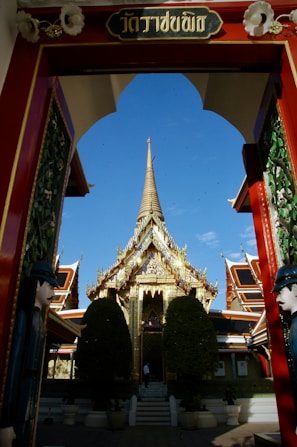 A grand, ornate temple entrance with a detailed archway leads to a glittering, golden temple structure. Intricate architectural details and lush greenery, including two large trees, are visible in the foreground. Vibrant colors and traditional design elements characterize the temple complex.