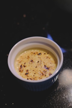 A cr&egrave;me br&ucirc;l&eacute;e dessert is placed in a white ramekin with a caramelized sugar crust on its surface. The background is a dark countertop, providing contrast to the light color of the dessert.