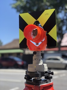 A close-up of a surveying instrument, specifically a total station, mounted on a tripod. The instrument features an orange cylindrical target with a reflective surface and a black and yellow square backing with a cross pattern. The background is blurred, showing some cars and trees, suggesting an outdoor setting.
