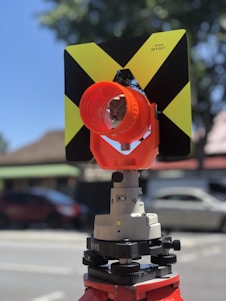 A close-up of a surveying instrument, specifically a total station, mounted on a tripod. The instrument features an orange cylindrical target with a reflective surface and a black and yellow square backing with a cross pattern. The background is blurred, showing some cars and trees, suggesting an outdoor setting.