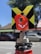 A close-up of a surveying instrument, specifically a total station, mounted on a tripod. The instrument features an orange cylindrical target with a reflective surface and a black and yellow square backing with a cross pattern. The background is blurred, showing some cars and trees, suggesting an outdoor setting.